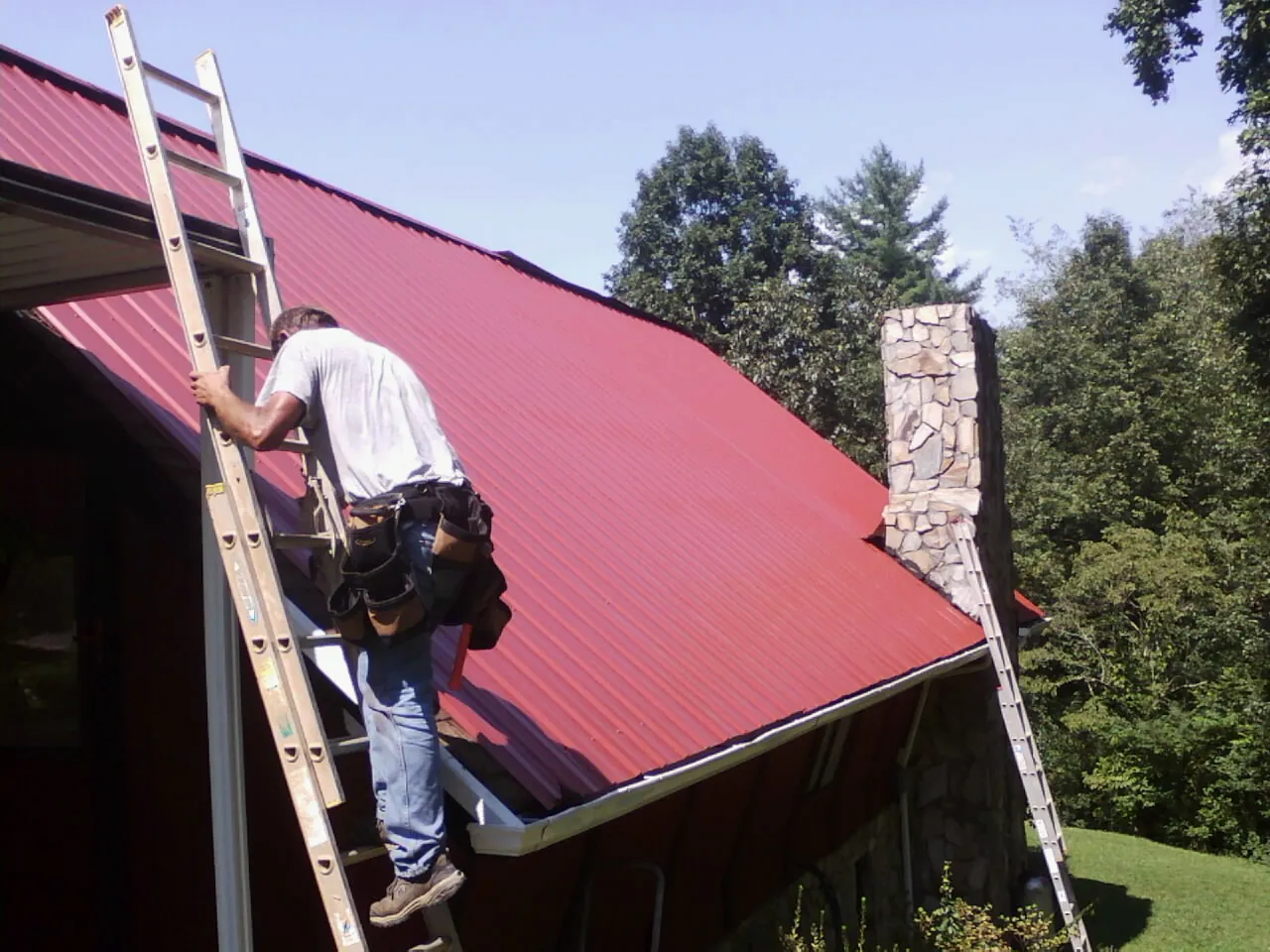 Roofer on a ladder beside a red metal roof and stone chimney — Soffit & Fascia Repair by Roofing Brothers in Pleasant Ridge